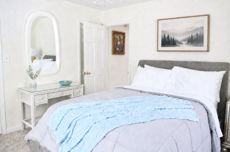 Amara Verde, the bed with a soft blue throw and a grey headboard, under a framed forest print.