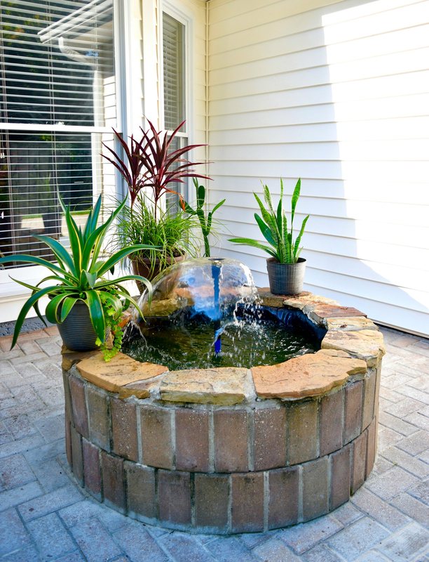 A round brick water fountain on the paved terrace at the front of the house.