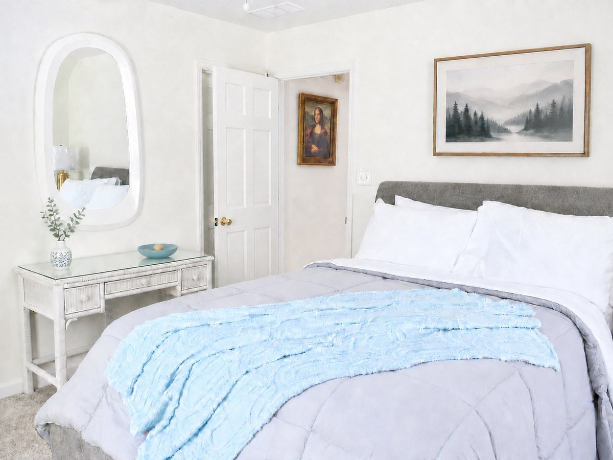Amara Verde Suite: the bed with its blue textured throw, a framed landscape above it, and the white vanity with an oval mirror and eucalyptus across the room.